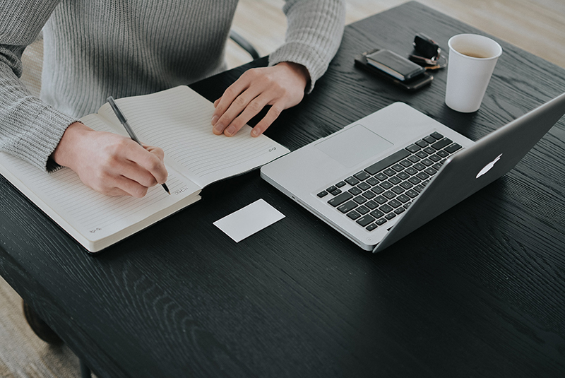 adult writing in a notebook in front of a laptop